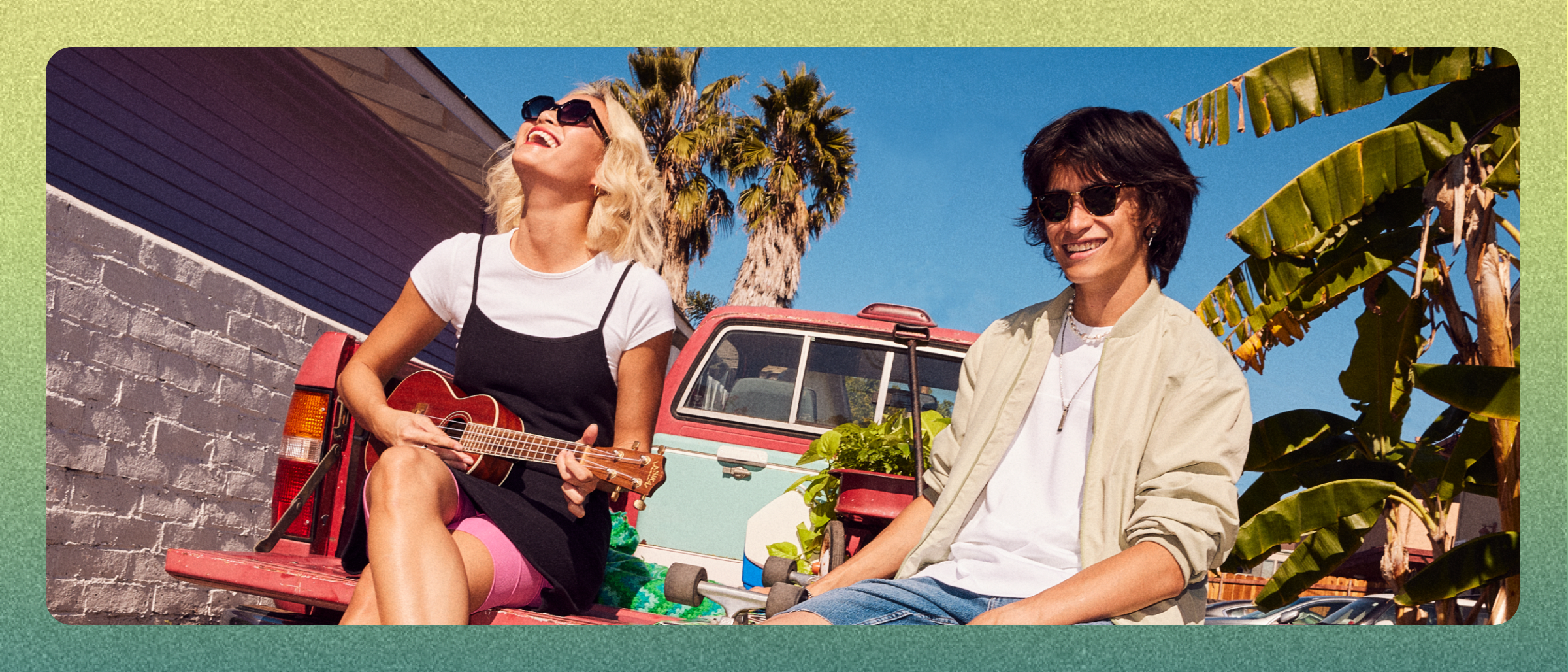 Two people sitting on the back of a red pickup truck smiling and playing ukelele
