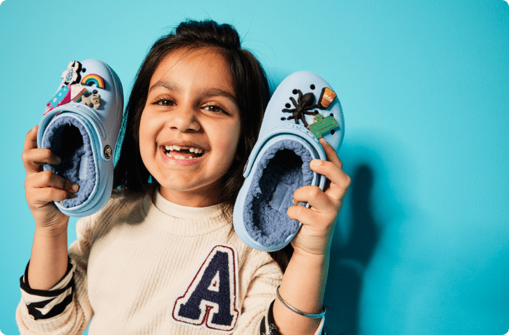 Child holding up fuzz-lined clogs filled with Jibbitz. Icon of box crossed out.