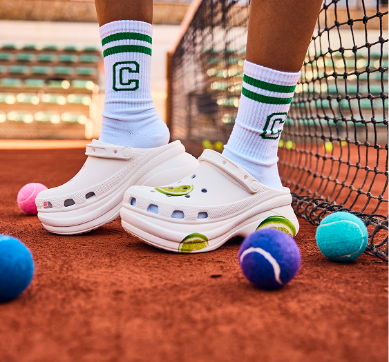 Legs of girl on tennis court, surrounded by tennis balls, wearing ankle socks and platform white crocs with limes on them