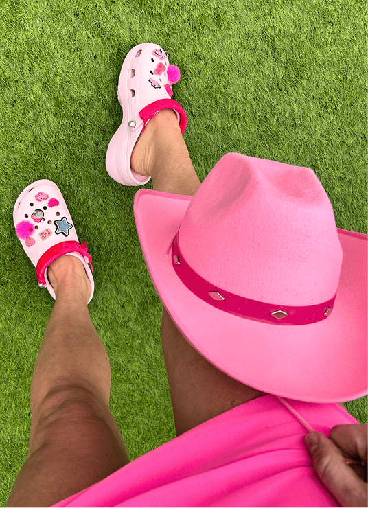 Legs of girl in hot pink skirt, holding a hot-pink cowgirl hat and wearing platform hot pink crocs with frilly pink jibbitz